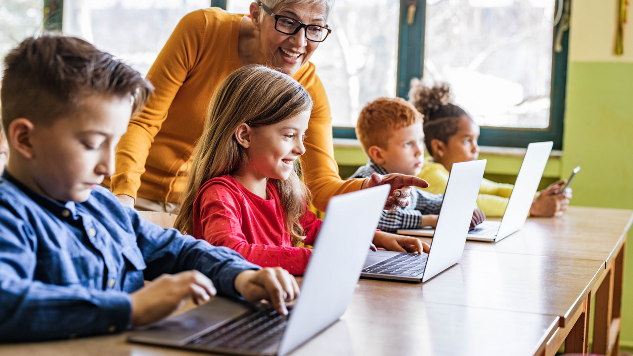 Mehrere Schülerinnen und Schüler die jeweils vor einem Laptop sitzen und arbeiten. Die Lehrerin steht hinter ihnen und hilft.