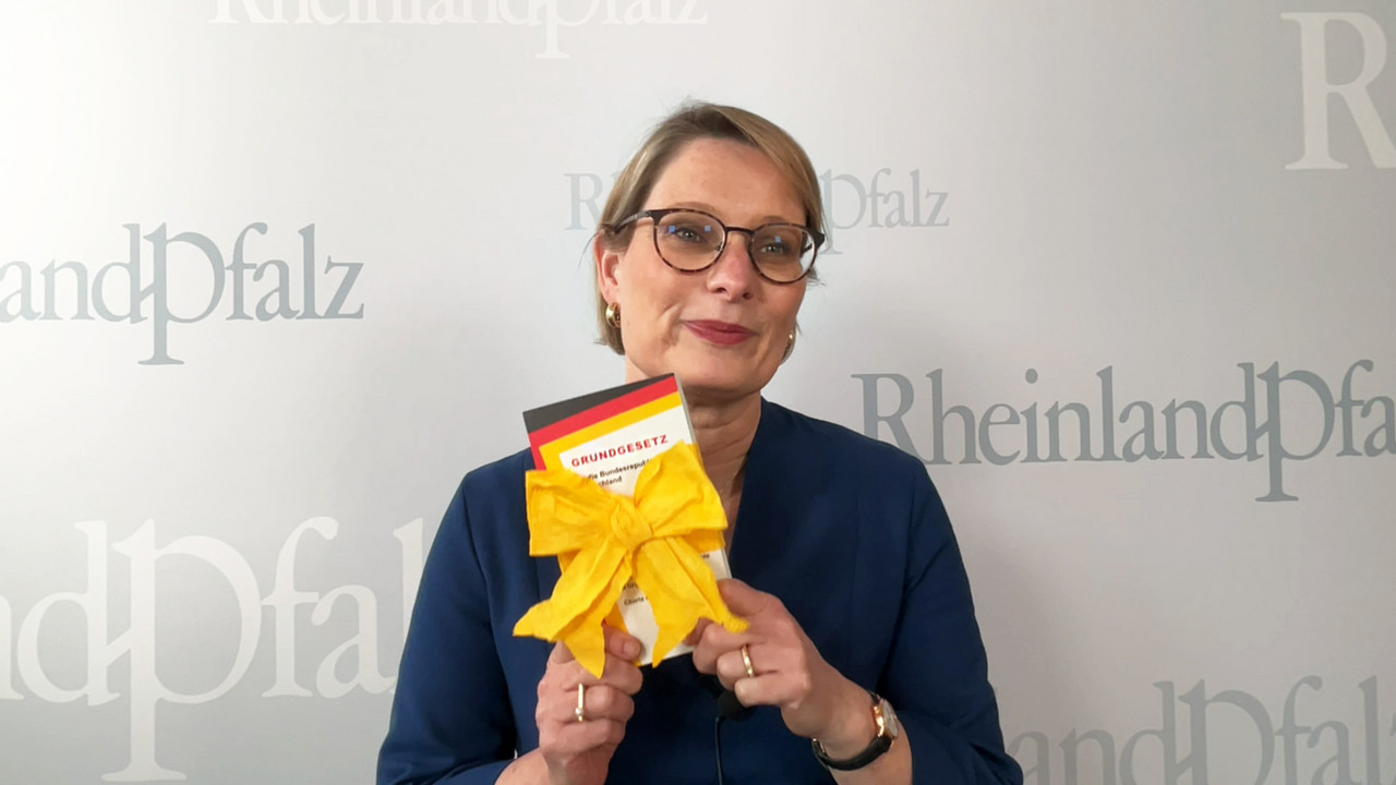 Zu sehen ist Bildungsministerin Dr. Stefanie Hubig. Sie hält ein mit Schleife umwickeltes Grundgesetz in die Kamera. 