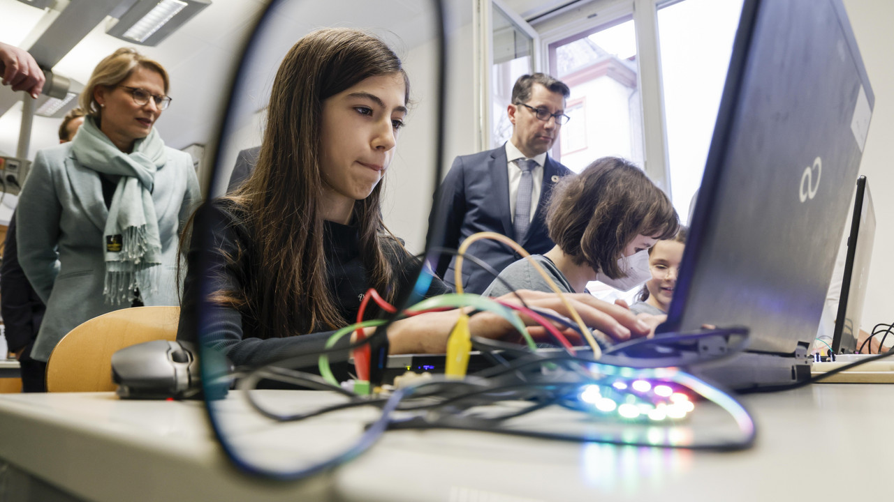 Eine Schülerin programmiert am Laptop eine Platine. Im Hintergrund steht Bildungsministerin Hubig und schaut der Schülerin zu. 